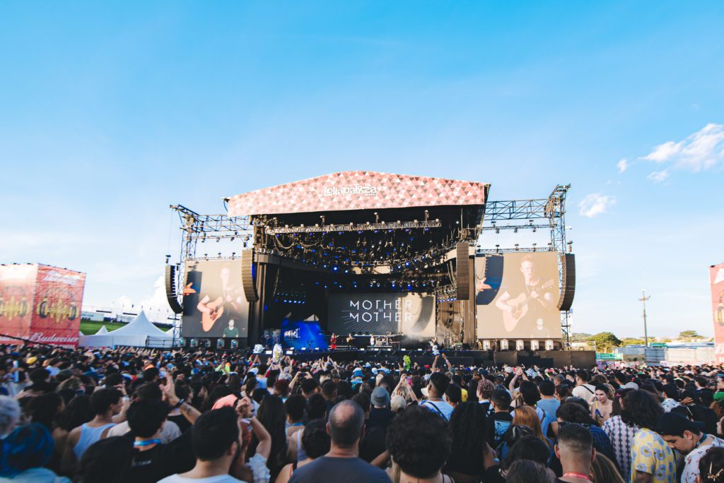 Palco de festival de música ao ar livre com a banda Mother Mother se apresentando para uma grande multidão sob céu azul.