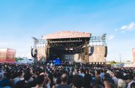 Palco de festival de música ao ar livre com a banda Mother Mother se apresentando para uma grande multidão sob céu azul.