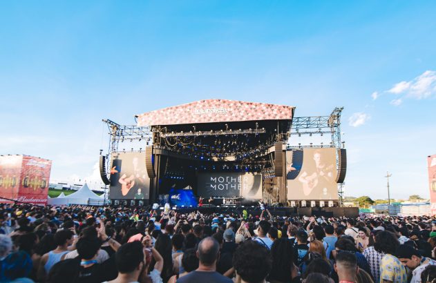 Palco de festival de música ao ar livre com a banda Mother Mother se apresentando para uma grande multidão sob céu azul.