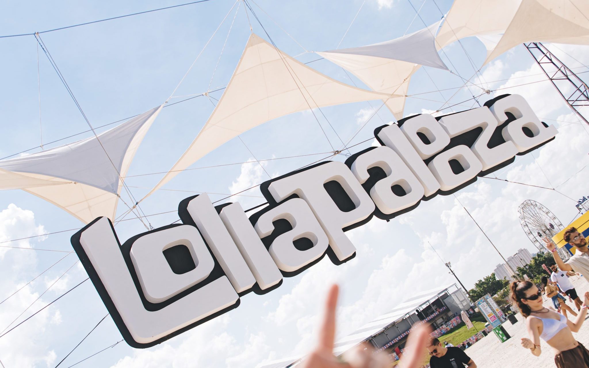 Letreiro Lollapalooza em festival com pessoas, céu azul, velas de sombra e roda gigante.