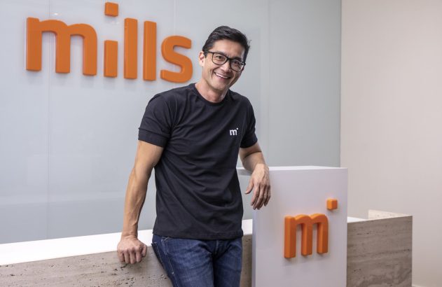 Homem sorridente de óculos e camiseta preta, encostado em balcão. Ao fundo, logo 3D 'mills'; no balcão, logo 'm'.