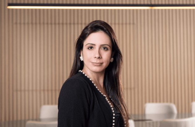 Mulher em ambiente de escritório, vestindo preto e pérolas, olhando para a câmera.