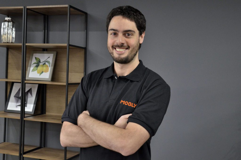 Homem sorridente de camisa polo preta Mobly, braços cruzados, em frente a estante decorada.