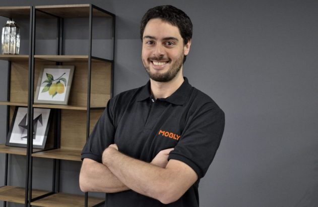 Homem sorridente de camisa polo preta Mobly, braços cruzados, em frente a estante decorada.