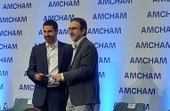 Homem entrega prêmio a outro em evento da AMCHAM no palco.