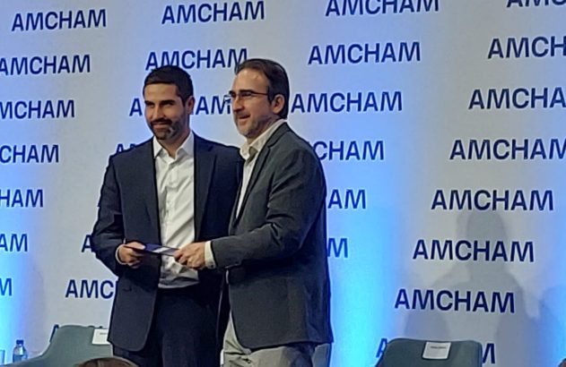 Homem entrega prêmio a outro em evento da AMCHAM no palco.