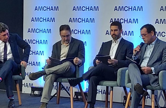 Evento AMCHAM com painel de quatro homens sentados; um com microfone, os demais com documentos.