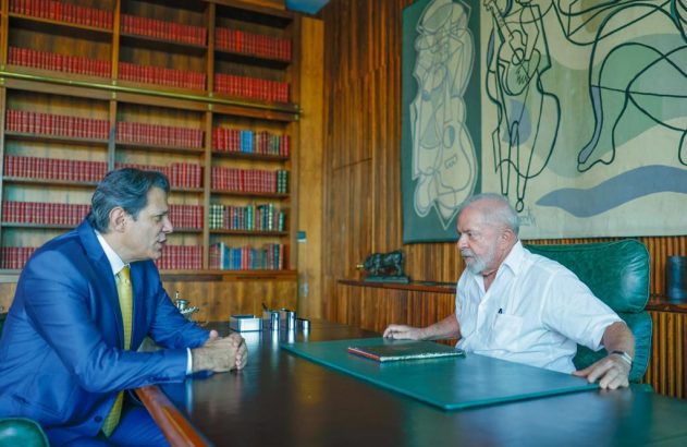 Lula e Haddad sentados à mesa em reunião. Ao fundo, estante de livros e tapeçaria abstrata.