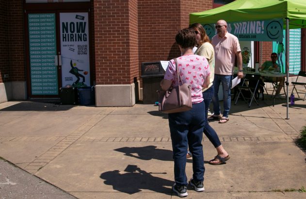Pessoas em calçada, próximas a loja Green District e tenda com sinalização de contratação.