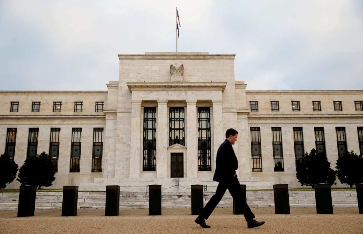 Homem de terno caminhando em frente ao edifício do Federal Reserve, com bandeira e selo da águia, sob céu nublado.