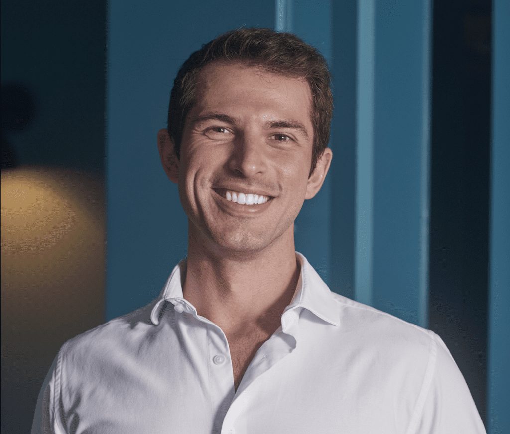 Retrato de homem jovem e sorridente, camisa branca, em frente a fundo azul com listras verticais.