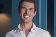 Retrato de homem jovem e sorridente, camisa branca, em frente a fundo azul com listras verticais.