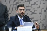 Homem de terno e óculos fala em microfone em audiência.