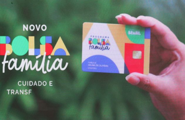 Mão segura cartão do Novo Bolsa Família exibido em tela. Destaques: logotipo e textos 'NOVO BOLSA família' e 'CUIDADO E TRANSF'.