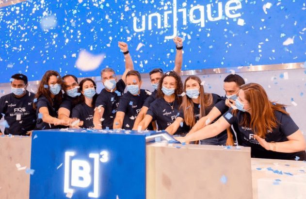 Equipe mascarada celebra lançamento com confetes e braços erguidos, apertando botões em pódio com logo B³ e Unifique.
