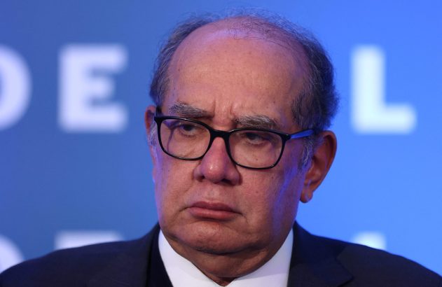 Homem de óculos, terno, com expressão pensativa. Fundo azul com letras.