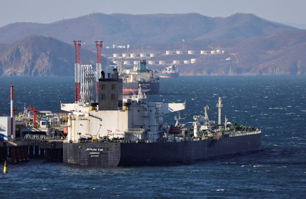Dois navios em porto industrial com guindastes e montanhas ao fundo.