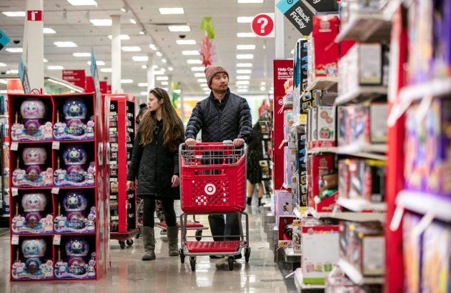 Clientes em um corredor da Target. Um homem empurra um carrinho vermelho enquanto uma mulher caminha ao lado, entre prateleiras de produtos.