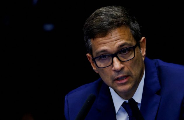 Homem de óculos e terno azul falando em microfones com fundo escuro.