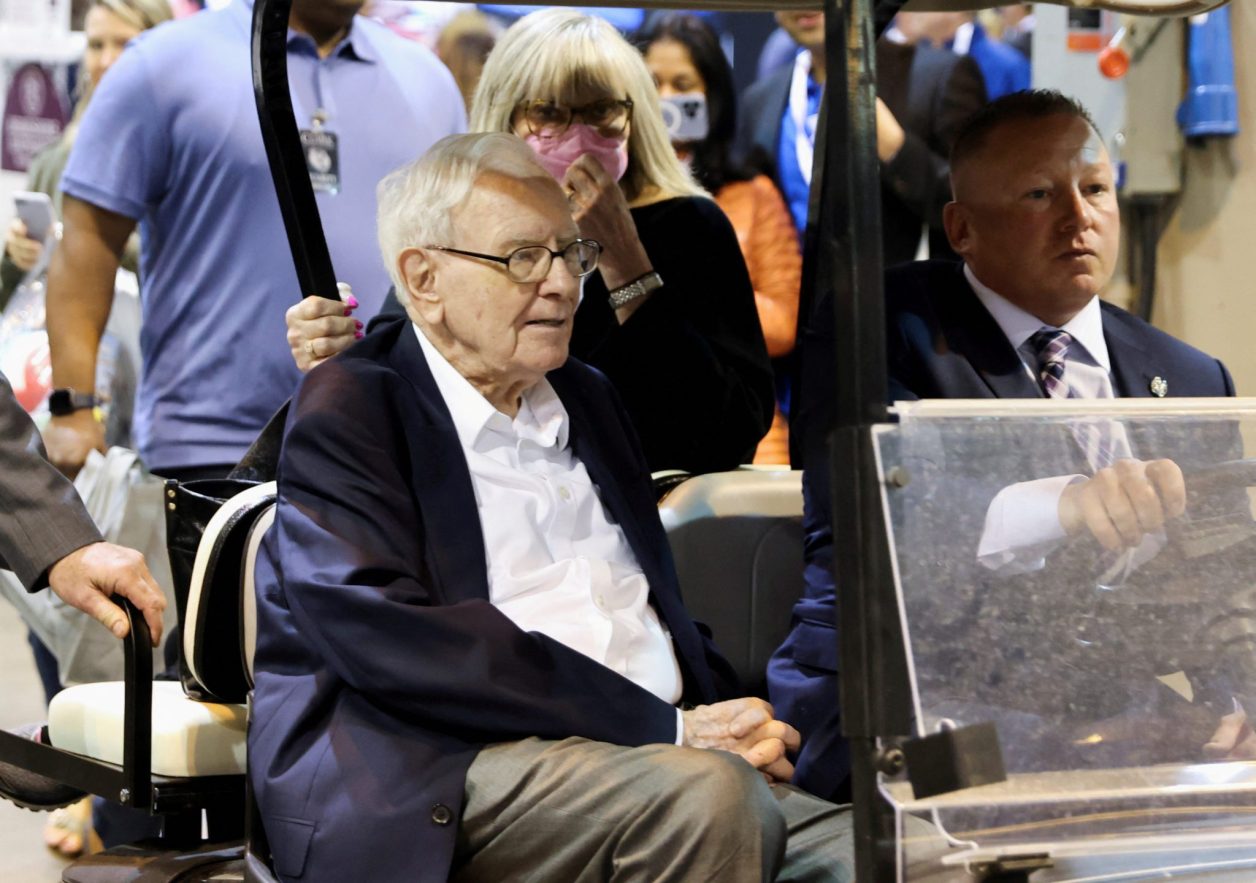Warren Buffett em carrinho de golfe, com motorista e mulher de máscara ao fundo.