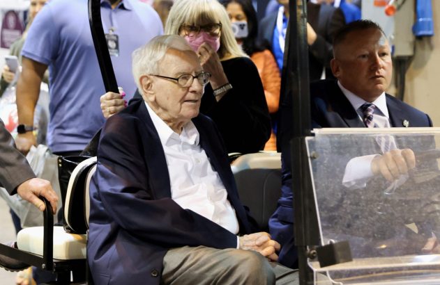 Warren Buffett em carrinho de golfe, com motorista e mulher de máscara ao fundo.