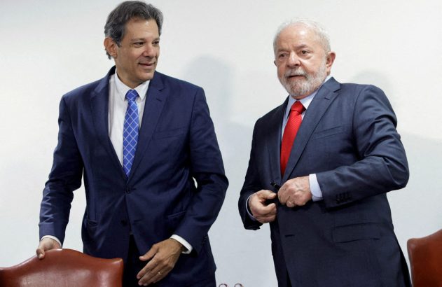Fernando Haddad sorri ao lado de Lula, de terno escuro e gravata vermelha, ajeitando o paletó.