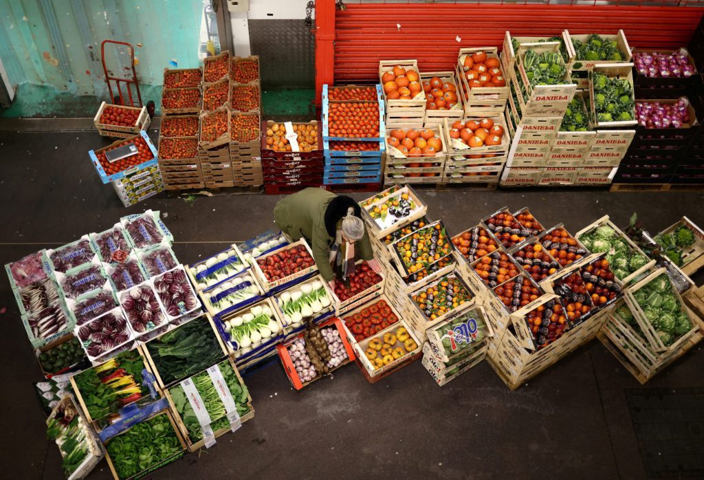 Mercado com frutas e vegetais frescos organizados em caixas de madeira no chão. Uma pessoa ao fundo.