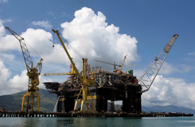 Plataforma de petróleo flutuante com guindastes, jorrando água, em estaleiro com montanhas e céu azul.