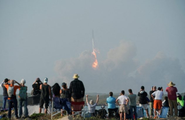 Grupo de pessoas assiste ao lançamento de um foguete ao longe, com fumaça e chamas intensas.