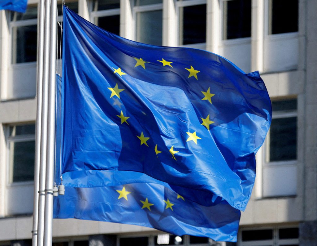 Bandeira azul da União Europeia com estrelas amarelas, tremulando contra um edifício claro.