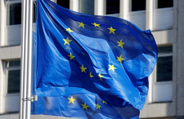 Bandeira azul da União Europeia com estrelas amarelas, tremulando contra um edifício claro.