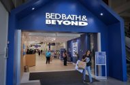 Entrada da loja Bed Bath & Beyond com pessoas. Mulher sai com sacolas.