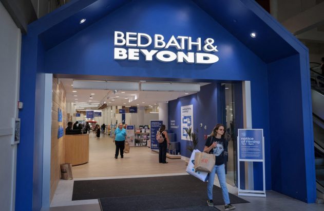 Entrada da loja Bed Bath & Beyond com pessoas. Mulher sai com sacolas.