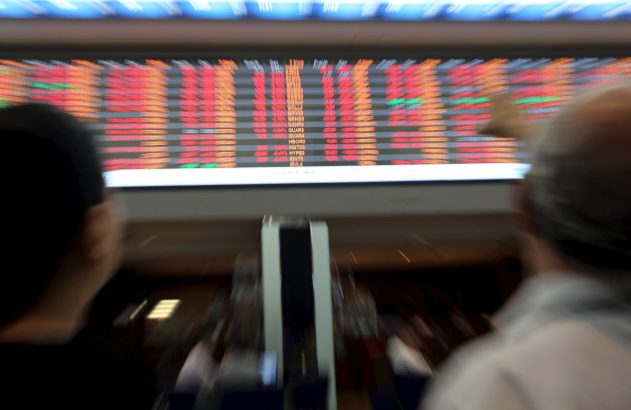 Vista de costas de duas pessoas observando um telão com cotações da bolsa de valores, muitas em vermelho, com borrão.