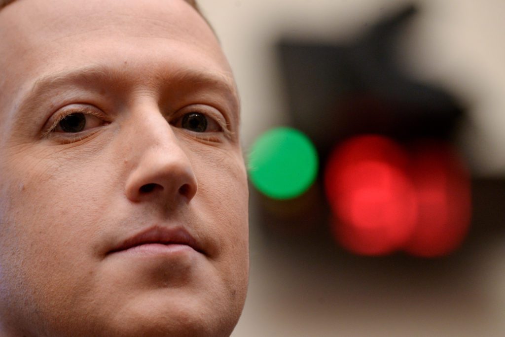 Mark Zuckerberg em close-up, olhando fixamente. Fundo desfocado com luzes verdes e vermelhas.