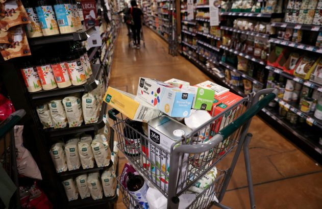 Carrinho de compras cheio de mantimentos em corredor de supermercado, com produtos nas prateleiras.