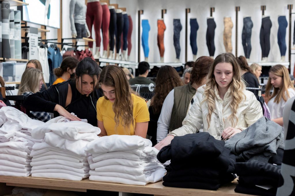 Clientes em uma loja de roupas examinam pilhas de peças dobradas sobre uma mesa. Mannequins com leggings ao fundo.
