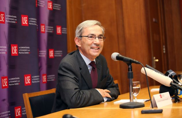 Homem de óculos e terno sorri em mesa com microfone e fundo LSE.