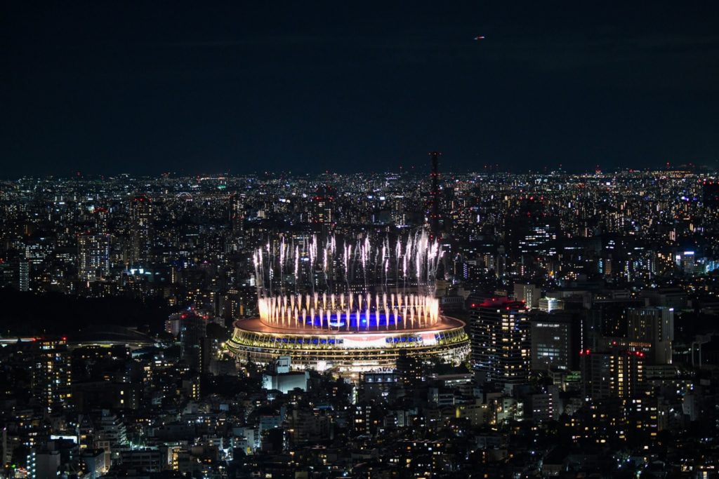Estádio iluminado à noite com fogos de artifício no céu, cercado por luzes da cidade. Vista aérea de evento.
