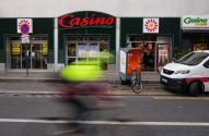 Ciclista borrado em movimento passa em frente a um supermercado Casino. Rua urbana com carro e bicicleta estacionados.