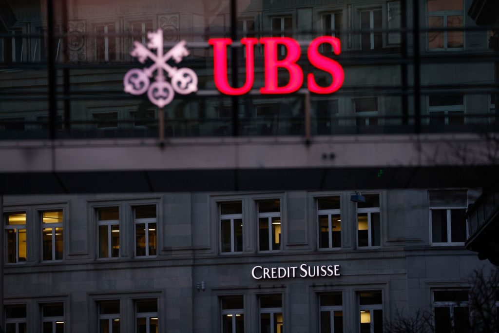 Placa iluminada da UBS acima da placa do Credit Suisse em um prédio à noite.