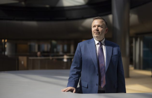Homem de terno azul, com barba, olha para o lado, com mão apoiada em mesa de ambiente moderno.