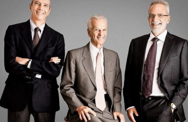 Três homens de terno sorrindo. Um em pé com braços cruzados, um sentado, e outro em pé com a mão no bolso.