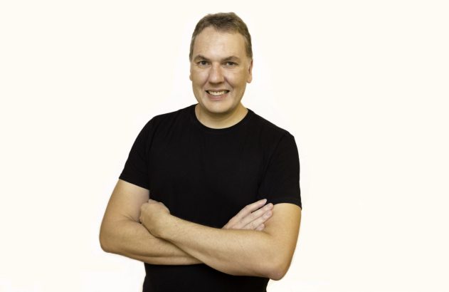 Retrato de um homem sorrindo, de braços cruzados, vestindo camiseta preta, contra fundo branco.