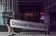 Dois homens carregam a placa "LEHMAN BROTHERS" em frente a um prédio. Um deles cobre o rosto.
