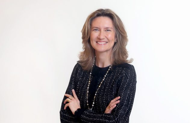 Mulher sorrindo, de braços cruzados. Veste blazer escuro e colar longo, em fundo branco.