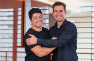 Dois homens sorrindo, um com o braço cruzado e o outro o abraçando pelo ombro, em ambiente interno.