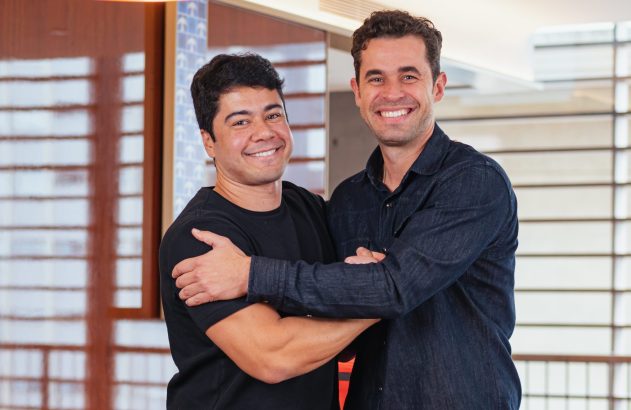 Dois homens sorrindo, um com o braço cruzado e o outro o abraçando pelo ombro, em ambiente interno.