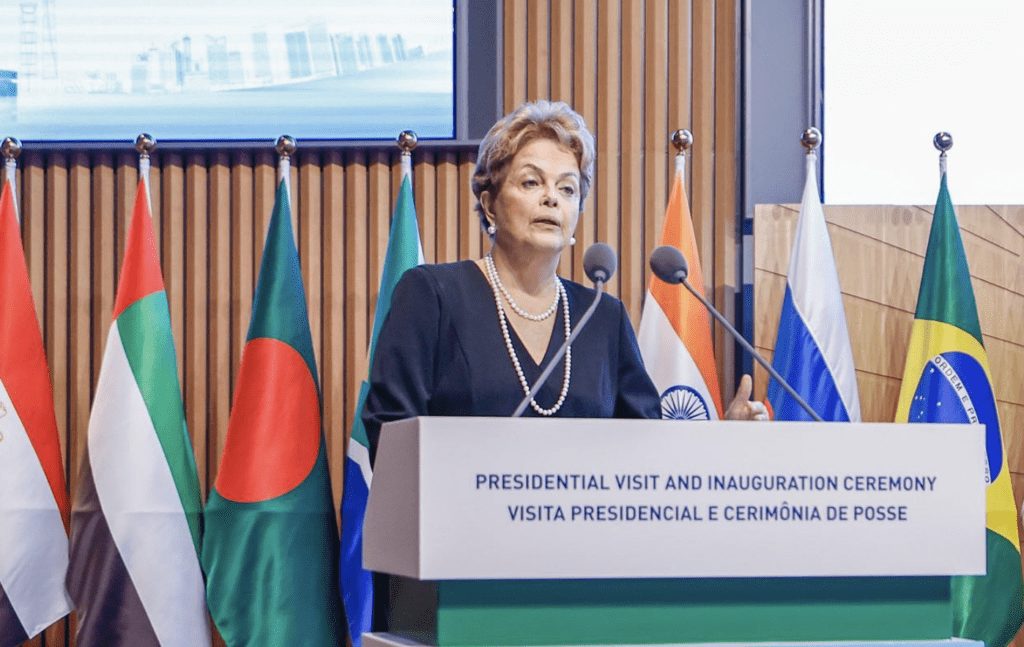 Mulher discursa em pódio, com bandeiras de nações ao fundo.