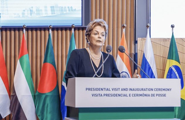 Mulher discursa em pódio, com bandeiras de nações ao fundo.